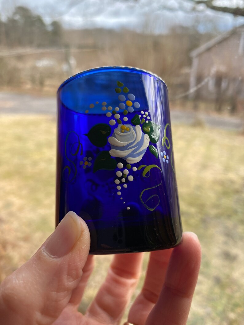 May include: A cobalt blue glass shot glass with a hand-painted floral design. The design features a white rose with green leaves and white dots.