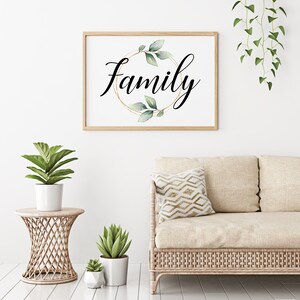 May include: A framed print with the word "Family" in elegant script, encircled by a gold ring and green leaves. The artwork is displayed above a wicker sofa with a patterned pillow, complemented by potted plants on a side table.