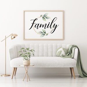 May include: A framed print with the word "Family" in elegant script, encircled by a gold ring and green leaves. The artwork hangs above a white sofa with a green throw and decorative pillow. A gold floor lamp and side table complete the scene.