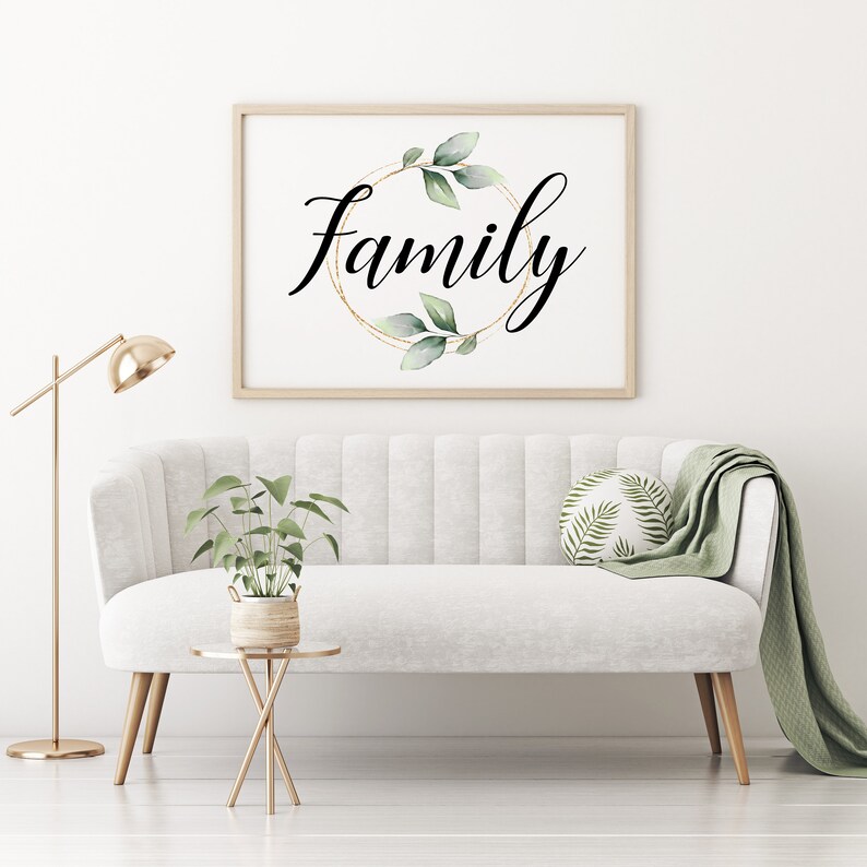 May include: A framed print with the word "Family" in elegant script, encircled by a gold ring and green leaves. The artwork hangs above a white sofa with a green throw and decorative pillow. A gold floor lamp and side table complete the scene.