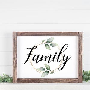 May include: A framed sign with the word "Family" in elegant black script. The word is centered within a gold-colored circular design, adorned with green leaves. The frame is made of dark brown wood, creating a rustic aesthetic.