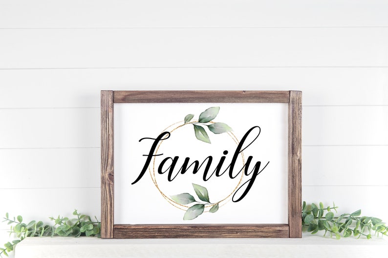 May include: A framed sign with the word "Family" in elegant black script. The word is centered within a gold-colored circular design, adorned with green leaves. The frame is made of dark brown wood, creating a rustic aesthetic.