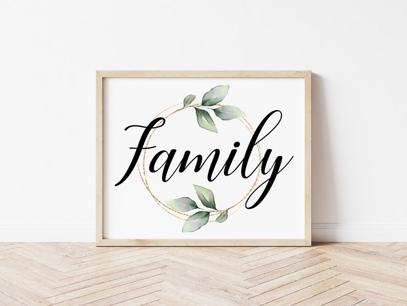May include: A framed print with the word "Family" in elegant black script. The word is centered within a gold-colored circular design, adorned with green and gray watercolor leaves. The frame is a light wood color.