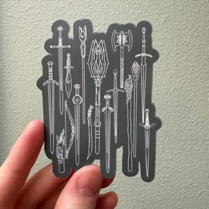Könnte beinhalten: Eine schwarz-weiße Aufkleberfolie mit einer Sammlung von 15 verschiedenen Schwert- und Waffendesigns. Die Aufkleberfolie befindet sich auf einem grauen Hintergrund.