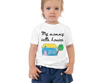 May include: White toddler t-shirt with the text "My mommy sells houses" in black script. Below the text is a colorful drawing of a house with a blue roof, yellow roof, pink door, and a green tree. The toddler is wearing blue shorts.