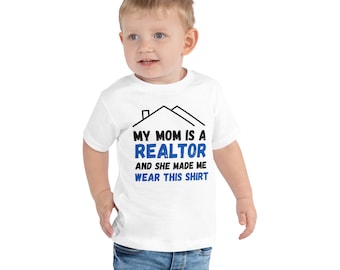 May include: White toddler t-shirt with black text that reads "My Mom is a Realtor and she made me wear this shirt". The text is surrounded by a black house outline.