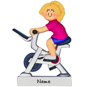 May include: A white and silver spin bike with a blonde figurine riding it. The figurine is wearing a pink shirt and blue shorts. The bike has a silver base with the word "Name" printed on it.