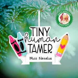 May include: A white Christmas ornament with the text "TINY HUMAN TAMER" and "Miss Natalie". The ornament features a pink and blue crayon, and a yellow pencil with a pink heart. A Santa Claus seal is also visible.