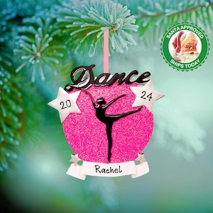 May include: Pink glitter Christmas ornament with a black silhouette of a dancer and the word "Dance" in black script. The ornament also has two silver stars with the numbers "20" and "24" on them. A white banner at the bottom says "Rachel".