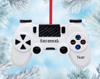 May include: A white video game controller ornament featuring black buttons, a black central touchpad, and colorful action symbols. The name Savannah is printed in black text above the center, with the word Year printed below the right buttons.