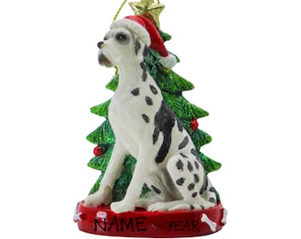 May include: A Christmas ornament featuring a white and black Dalmatian dog wearing a red Santa hat, sitting in front of a green Christmas tree. The ornament has a red base with the words "NAME YEAR" on it.