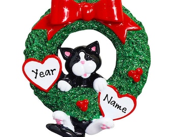 May include: A Christmas ornament featuring a black and white cat inside a green wreath with a red bow. The cat holds two heart-shaped signs, one with the word "Year" and the other with the word "Name". Red berries are also included.