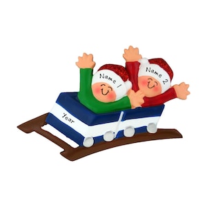 May include: Personalized Christmas ornament featuring two children in a blue and white sled on a brown track. The children are wearing red and green Santa hats and have smiling faces. The ornament includes the text "Name 1", "Name 2", and "Year".