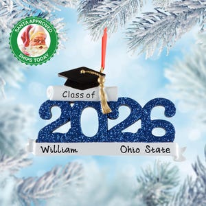 May include: A graduation-themed Christmas ornament featuring the blue glittery numbers 2026, a black mortarboard cap, and a white diploma scroll. The white banner at the bottom displays the text William and Ohio State in black lettering.