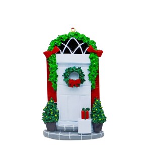 May include: A festive Christmas ornament featuring a white door with a green wreath and red bow. The door is set within a red frame, decorated with green garland and red bows. Two small Christmas trees and a gift box are also present.