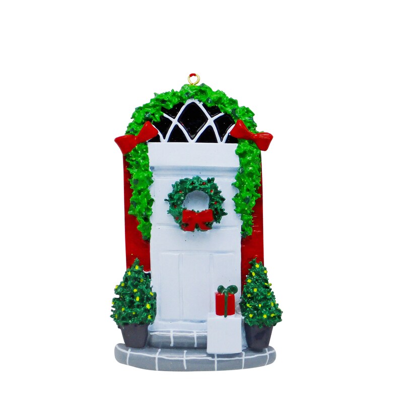 May include: A festive Christmas ornament featuring a white door with a green wreath and red bow. The door is set within a red frame, decorated with green garland and red bows. Two small Christmas trees and a gift box are also present.