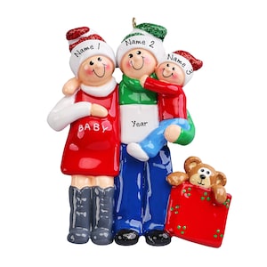May include: A family Christmas ornament featuring a pregnant woman, a man, and a child. The woman wears a red dress with "BABY" printed on it. The man holds the child, and a teddy bear peeks out of a red gift box. Each figure wears a hat with a name.