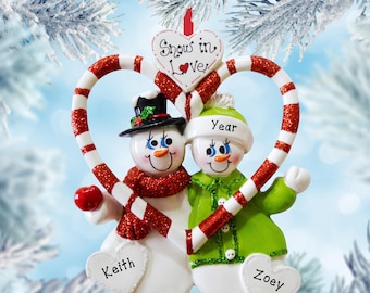 May include: A Christmas ornament featuring two snowmen framed by a red and white glittery candy cane heart. The top heart tag reads "Snow in Love". The left snowman wears a black top hat and red scarf, labeled "Keith". The right snowman wears a green hat and coat, labeled "Year" and "Zoey".