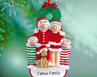 May include: A Christmas tree ornament shaped like a family of four and a dog. The figures wear red and white holiday clothing, with names Ryan, Kimberly, Kayls, Kylan, and Lola printed on hats. A green wreath sits at the top, and James Family is at the base.