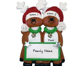 May include: Personalized Christmas ornament featuring two reindeer wearing Santa hats and holding a sign with the text "Family Name" and "Year".