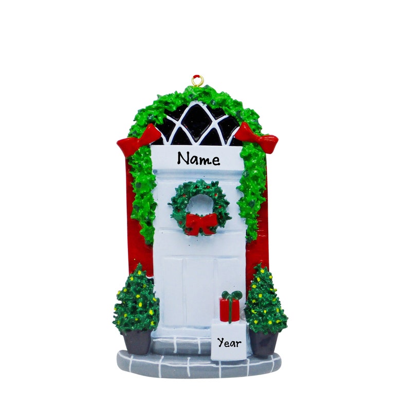May include: A festive Christmas door ornament featuring a white door with red trim, adorned with a green wreath and garland. The ornament includes red bows, two small Christmas trees, and the words "Name" and "Year".