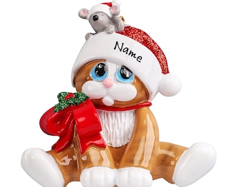 May include: A brown and white cat wearing a red and white Santa hat with a white pom pom. The cat is holding a red bow with green holly leaves. The hat has the word "Name" written on it. A small gray mouse is sitting on top of the hat.
