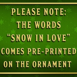 May include: A green background with gold snowflakes and a gold border. The text "PLEASE NOTE: THE WORDS "SNOW IN LOVE" COMES PRE-PRINTED ON THE ORNAMENT" is written in gold letters.
