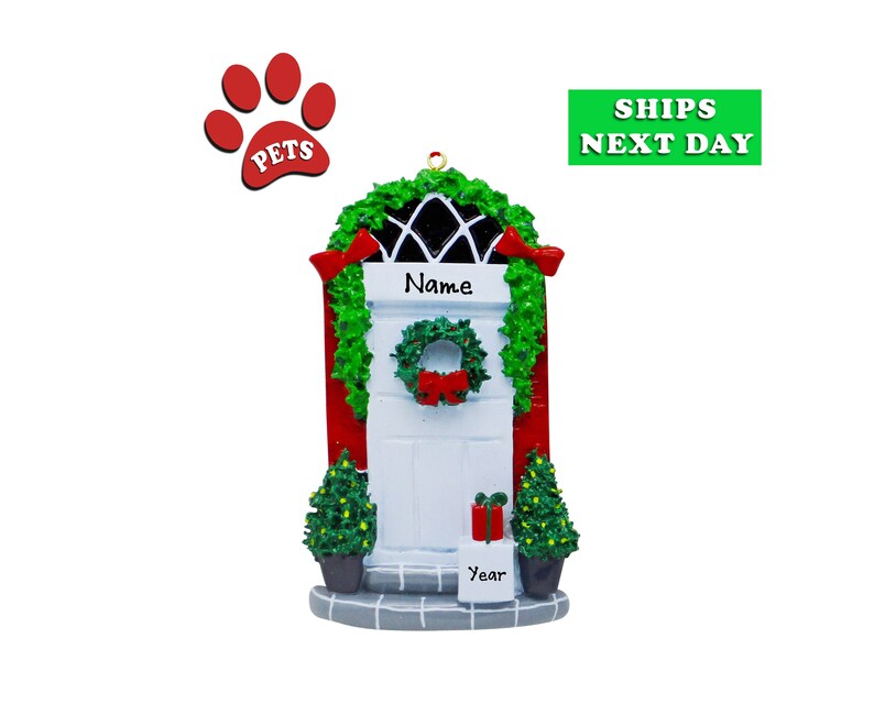 May include: A white Christmas door ornament with a green wreath and red bow. The door is trimmed in red and has a small Christmas tree on each side. The ornament has the word "Name" above the door and "Year" below. A red paw print with the word "PETS" is above the door.