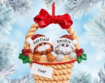 May include: A Christmas ornament shaped like a woven basket containing two kittens wearing red glittery Santa hats. The left kitten is orange with stripes and labeled Garfield, while the right is gray and labeled Smokie. A red bow sits on top, with a white tag labeled Year.