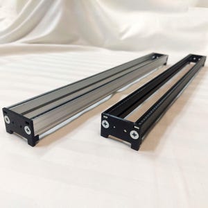 Puede incluir: Dos marcos rectangulares largos y estrechos de extrusión de aluminio, uno con acabado plateado metálico y otro con acabado negro mate. Cada marco cuenta con tapas de plástico negro aseguradas por dos tornillos de cabeza Phillips plateados.