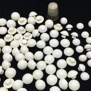 Può includere: Un grande mucchio di bottoni di plastica bianca con un piccolo ditale d'argento sullo sfondo.