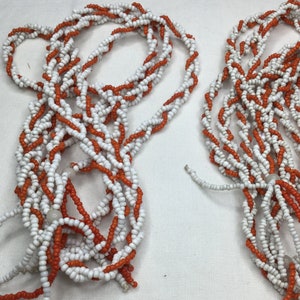 Peut inclure: Deux colliers de perles, chacun composé de perles blanches et orange. Les perles sont enfilées selon un motif torsadé, créant un design texturé et visuellement intéressant. Les colliers sont probablement faits à la main.