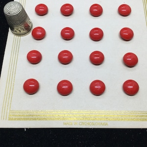 Puede incluir: Un conjunto de pequeños botones redondos de color rojo brillante dispuestos sobre una tarjeta crema con bordes dorados. Una dedal de plata descansa sobre la tarjeta. La tarjeta está etiquetada como "MADE IN CZECHOSLOVAKIA".