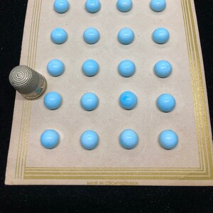 Peut inclure: Une carte avec 20 boutons ronds bleu clair disposés en grille. La carte a une bordure dorée et le texte "MADE IN CZECHOSLOVAKIA" en bas. Un dé à coudre argenté se trouve dans le coin inférieur gauche.