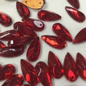 Könnte beinhalten: Eine Ansammlung von tropfenförmigen, facettierten, roten Glas-Strasssteinen. Jeder Stein hat ein spitzes Ende und ein Loch zum Auffädeln. Die Steine liegen auf einer weißen Oberfläche, mit ein paar goldfarbenen Teilen.
