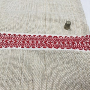 May include: A close-up of a white linen fabric with a red and white woven border. The fabric has a subtle texture and a natural, rustic look.