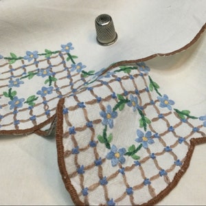 May include: A white linen tablecloth with a brown scalloped edge and embroidered blue flowers and brown latticework.