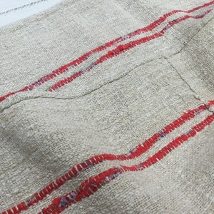 May include: A close-up of a piece of off-white fabric with two red stripes running horizontally across the fabric. The fabric has a rough texture and appears to be woven.