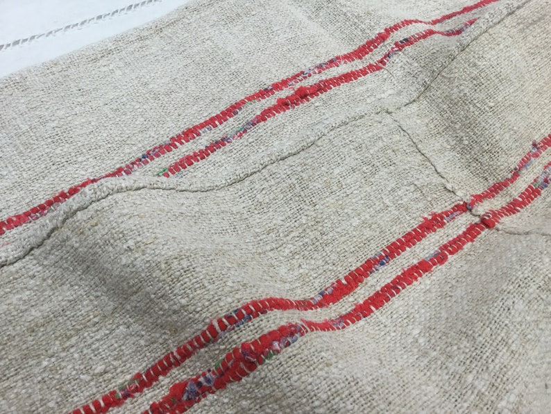 May include: A close-up of a piece of off-white fabric with two red stripes running horizontally across the fabric. The fabric has a rough texture and appears to be woven.