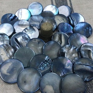 Peut inclure: Un ensemble de boutons en coquillage bleu-gris irisé avec un dé à coudre doré au centre. Les boutons ronds varient en couleur, certains reflétant des nuances de violet et de vert. Le dé à coudre est en matériau doré tissé.