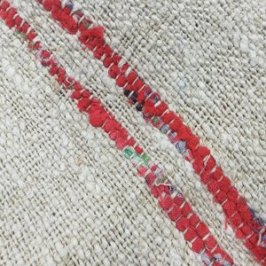 May include: Close-up of a woven rug with two red stripes running diagonally across the surface. The rug is made of a natural fiber and has a rough texture.