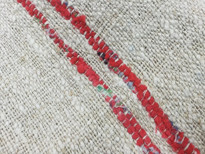 May include: Close-up of a woven rug with two red stripes running diagonally across the surface. The rug is made of a natural fiber and has a rough texture.