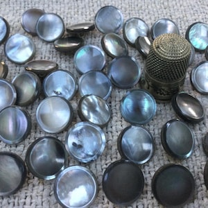 Può includere: Una collezione di bottoni rotondi iridescenti in tonalità di grigio, nero e argento, insieme a un ditale decorativo in metallo. I bottoni variano per dimensioni e lucentezza, suggerendo un tema vintage o artigianale.