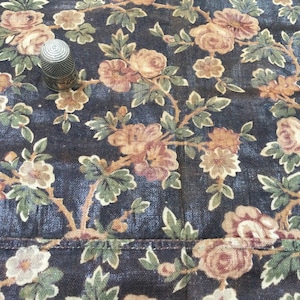 Könnte beinhalten: Ein dunkelblauer Stoff mit einem floralen Muster aus braunen und rosafarbenen Rosen und grünen Blättern. In der Mitte des Bildes befindet sich ein silberner Fingerhut.