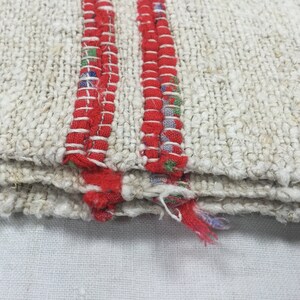 May include: Close-up of a white woven rug with two red stripes along the edge. The rug is folded in thirds, showing the layers of the weave.