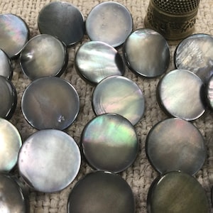 May include: A selection of iridescent, circular mother-of-pearl shell buttons in shades of grey and silver. The buttons are displayed on a textured, beige surface, with a silver thimble in the background. These buttons are suitable for sewing and craft projects.