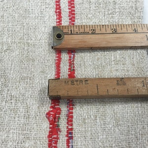 May include: A close-up of a white linen fabric with two red stripes. Two wooden rulers are placed on top of the fabric. The top ruler is marked in inches and the bottom ruler is marked in centimeters. The top ruler reads "Made in Canada" and the bottom ruler reads "M&egrave;tre".