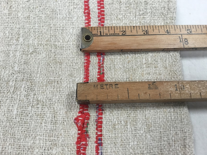 May include: A close-up of a white linen fabric with two red stripes. Two wooden rulers are placed on top of the fabric. The top ruler is marked in inches and the bottom ruler is marked in centimeters. The top ruler reads "Made in Canada" and the bottom ruler reads "M&egrave;tre".