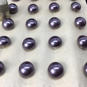 Peut inclure: Une collection de boutons ronds violets avec une finition nacrée. Les boutons sont disposés en rangées sur une surface de couleur claire. Un dé à coudre argenté est dans le coin supérieur gauche.