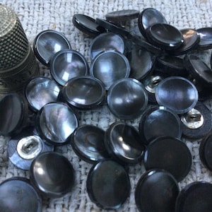 Op de afbeelding: Een verzameling zwarte, ronde knopen met een glanzende, parelmoerachtige glans. Er is ook een zilveren vingerhoed aanwezig. De knopen variëren in grootte en liggen verspreid op een getextureerd, crèmekleurig oppervlak. Deze knopen zijn ideaal voor naai- en knutselprojecten.